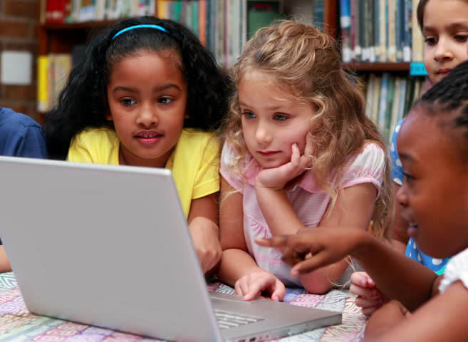 students under age 10 in front of a bookshelf reading from a laptop